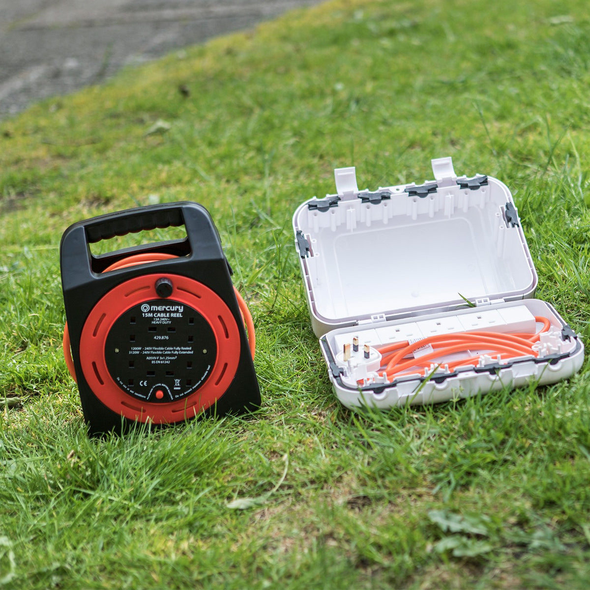 Mercury 15m 4-gang cable reel in red and black with orange cable, shown on grass beside an open white outdoor socket cover box.