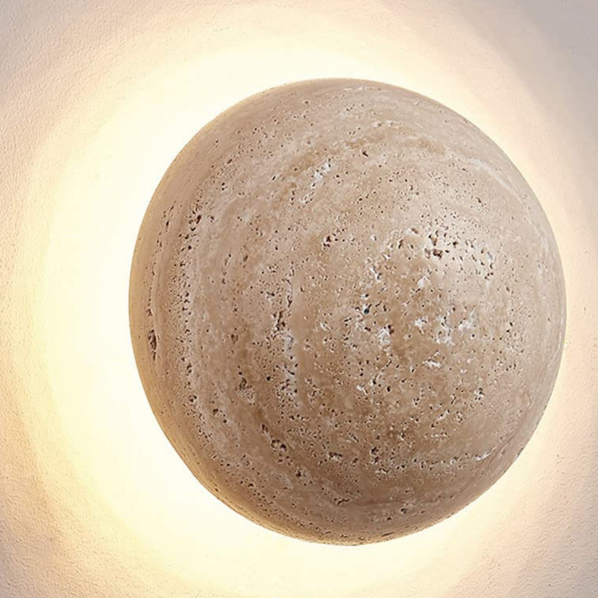 The Velora LED Spherical Travertine Wall Light in light beige casts a warm glow against a pale wall, creating a soft halo around its round, textured stone design.