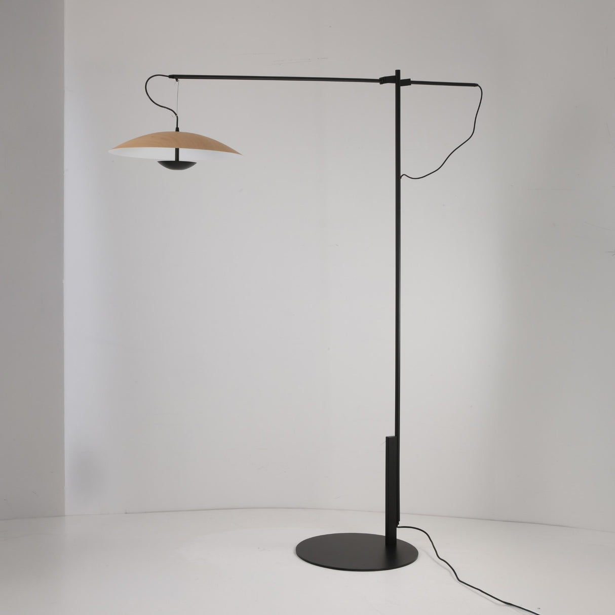 The Ginger LED Large Floor Lamp - Black & Brown features a black metal stand, circular base, and a horizontal arm with a wide, flat beige shade; its cord drapes along the arm and base, set against a plain white wall.