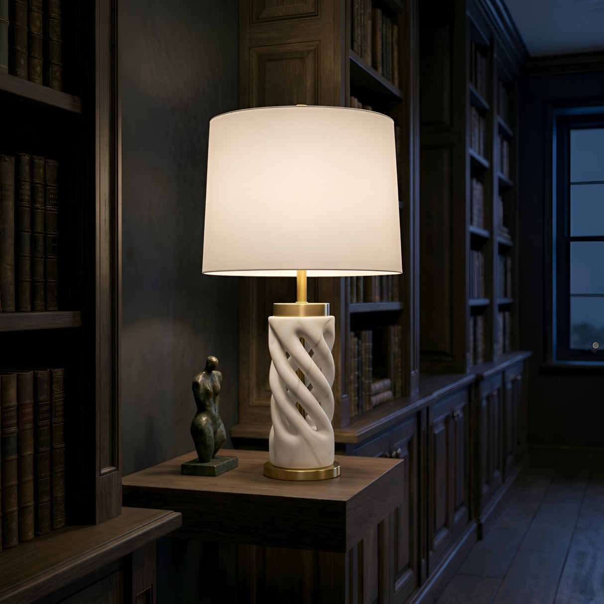 The Iverne 1 Light Twisted Carved Alabaster Table Lamp in Brass & Soft White sits on a wooden side table beside an abstract sculpture, framed by bookshelves in a dimly lit study.