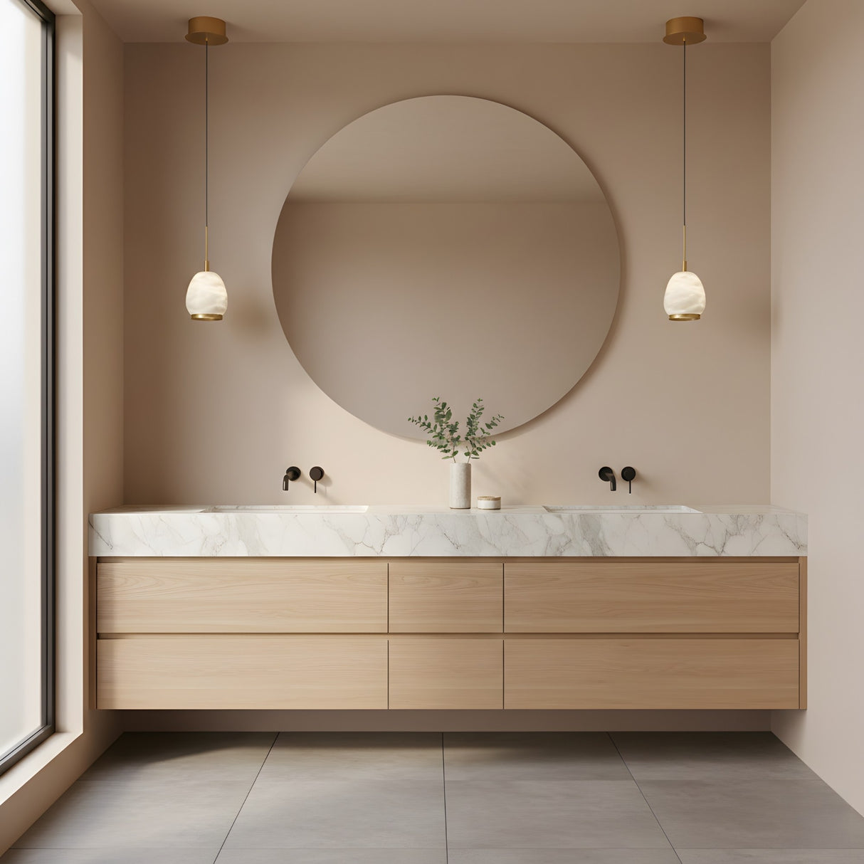 A modern bathroom with a large round mirror, light wood vanity with marble countertop, double sinks with black fixtures, and a vase of greenery. Natural light is enhanced by the Aurevia LED Drop Alabaster Single Pendant Light in brushed brass.