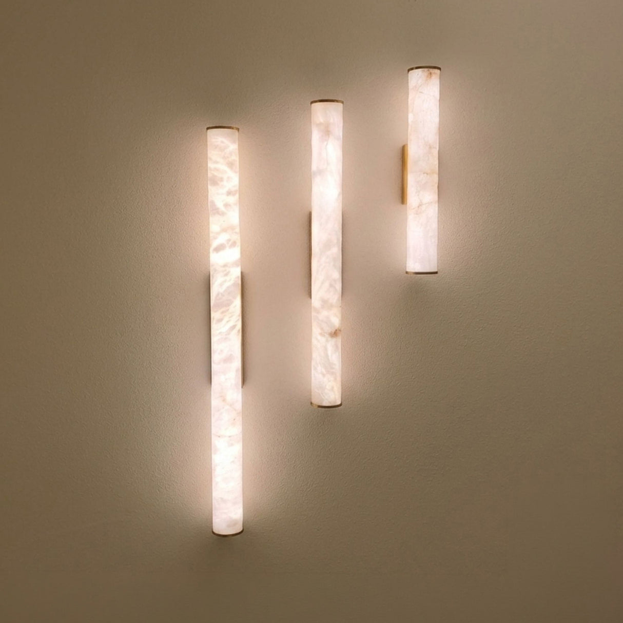 Montavo LED alabaster tube wall lights, 800mm cylinders in natural stone with brushed brass caps and backplates, mounted at varying heights, glowing warm white against a neutral wall.