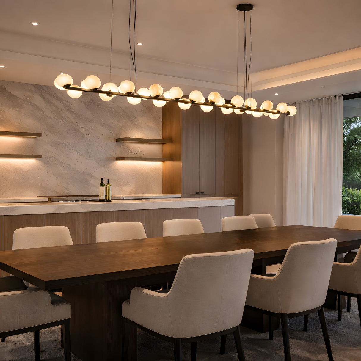 A modern dining room features a long wooden table, cream upholstered chairs, built-in shelves, and the Axis 27 Light Alabaster Suspension Light – Matte Black above. Marble backsplash and large windows with sheer curtains fill the space with natural light.