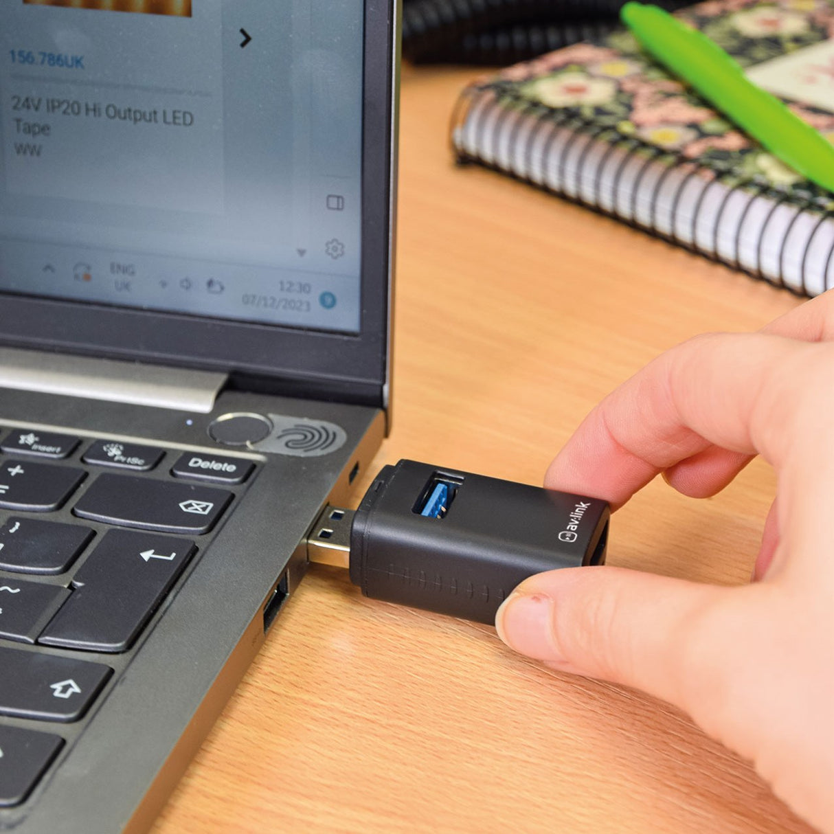 Hand plugging the AV:Link 3 Port Mini USB-A Hub into a laptop's USB port on a wooden desk, showing the matte black casing and USB 3.2 port.