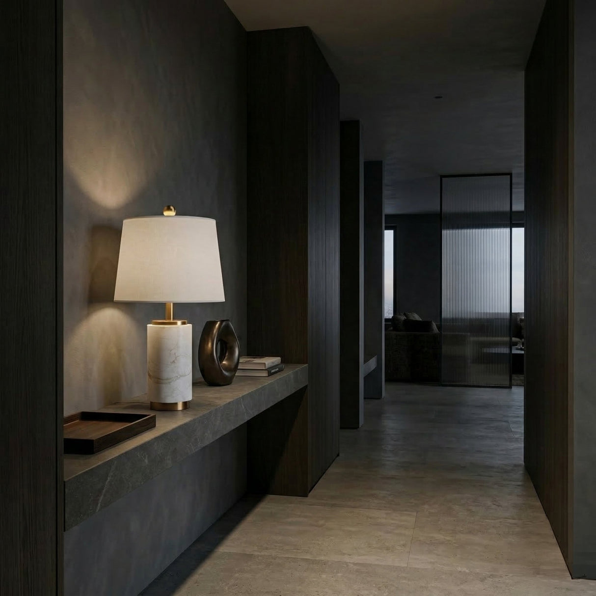 A modern, dimly lit hallway with dark walls and light stone flooring features a floating shelf displaying the Valmera 1 Light Cylinder Marble Table Lamp in brass and white, alongside a black sculpture and tray. In the background, a glass door leads to another room.