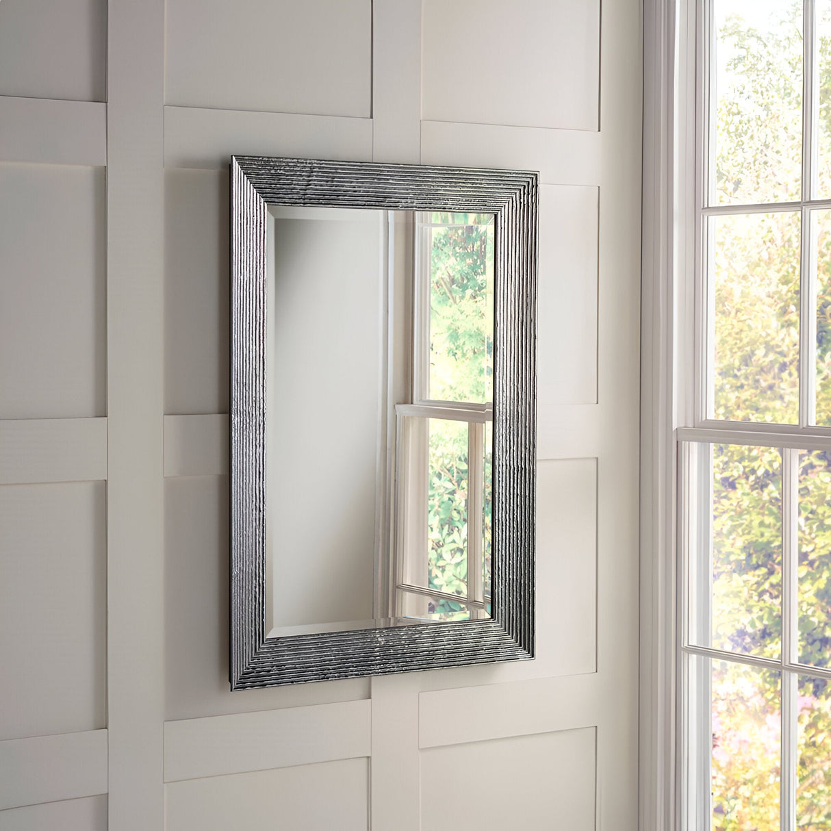 Contemporary elegance fills the room as the Charm Rectangle Wall Mirror in Silver, featuring a textured metallic frame, adorns the white paneled wall. Sunlight streams through the window on the right, casting reflections and revealing a view of lush green foliage outside.