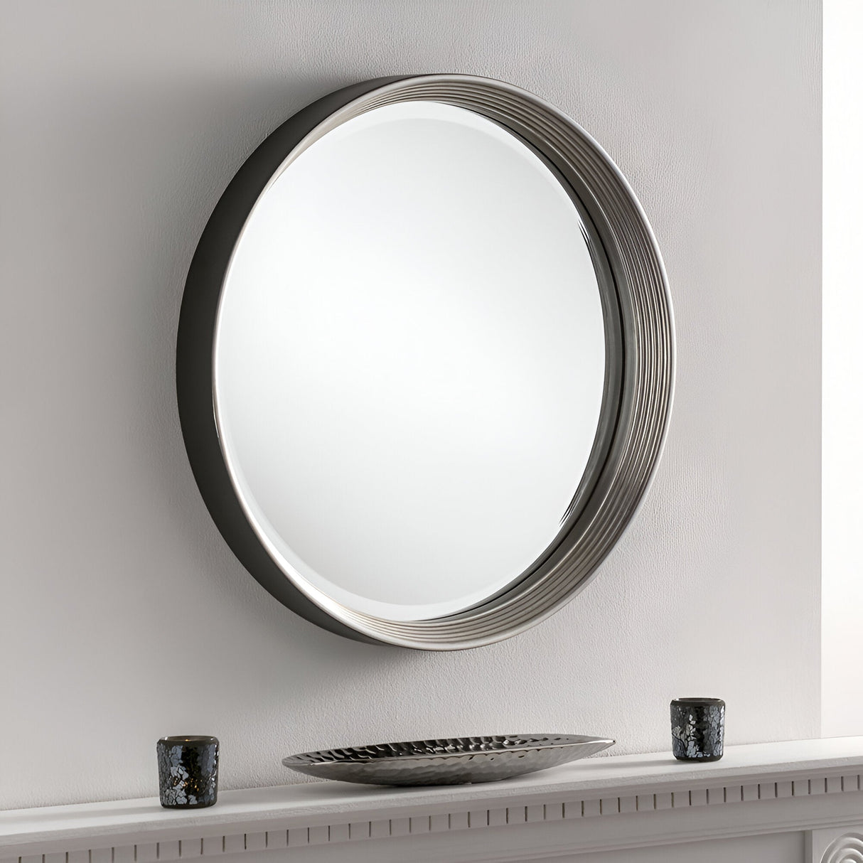 A Solune Circle Wall Mirror Black & Silver - 61cm is mounted on a white wall above a mantel, enhancing modern aesthetics. Two small candle holders and a decorative bowl complete the contemporary design below the mirror.