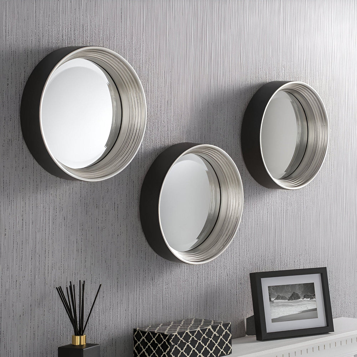 Three Solune Circle Wall Mirrors (61cm, black & silver) hang on a textured gray wall. Below them, a black and white photo rests on a table with a decorative black box and reed diffuser, creating sleek modern aesthetics.