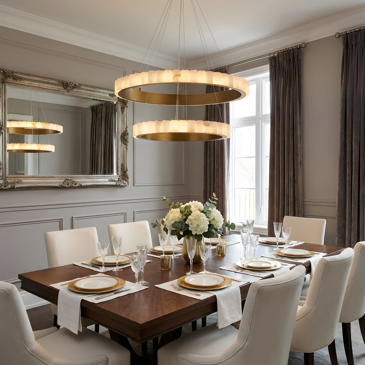 A modern dining room features a wooden table for six, white chairs, a large mirror, gray curtains, neutral walls, and elegant lighting with the Lucenza LED 2 Tier Halo Ring Alabaster Chandelier in Brushed Brass & Soft White above a white floral centerpiece.