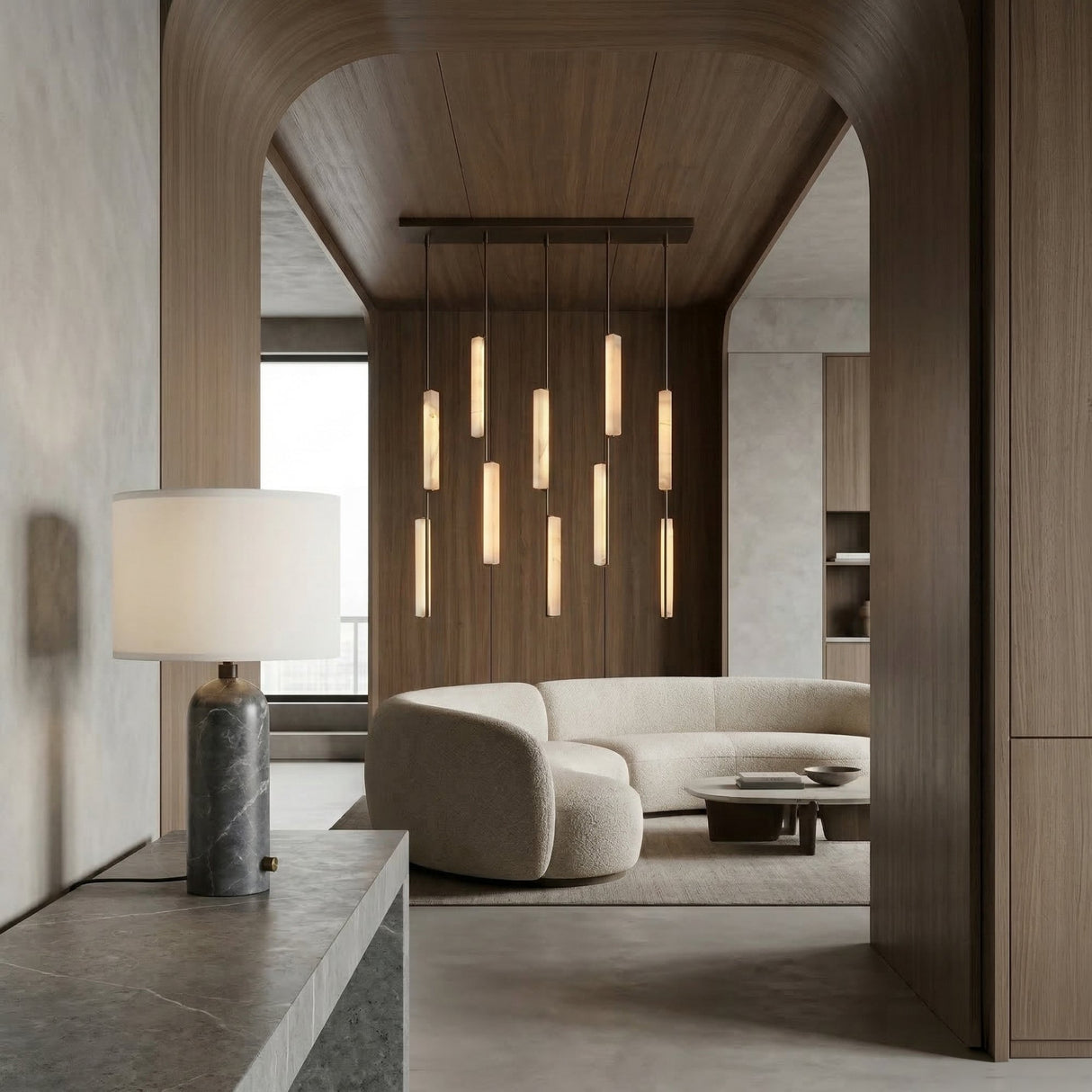 A modern living room features a curved cream sofa, round coffee table, pendant lights, and wooden walls. In the foreground, the Arvello 1 Light Minimal Marble Table Lamp - White & Grey sits on a stone console beneath an arched doorway.