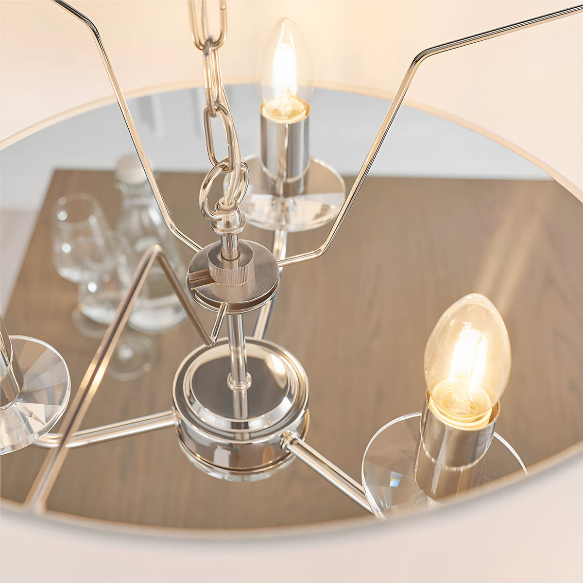 Close-up of Endon Castdon 3 light pendant in nickel with lit E14 candle bulbs, K9 crystal bobeches, chain suspension and mirrored disc above a dining table.