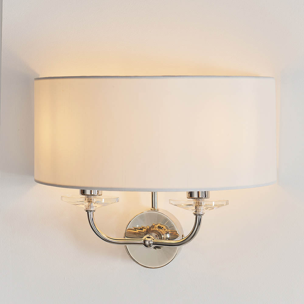 Castdon 2 Light Wall Light in nickel with white semi-circular fabric shade, twin arms and crystal accents, shown on a plain wall background.