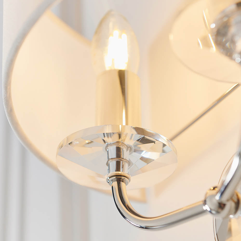Close-up of the Castdon 2 Light Wall Light in nickel, showing a lit E14 bulb, faceted crystal disc detail and white faux silk shade.