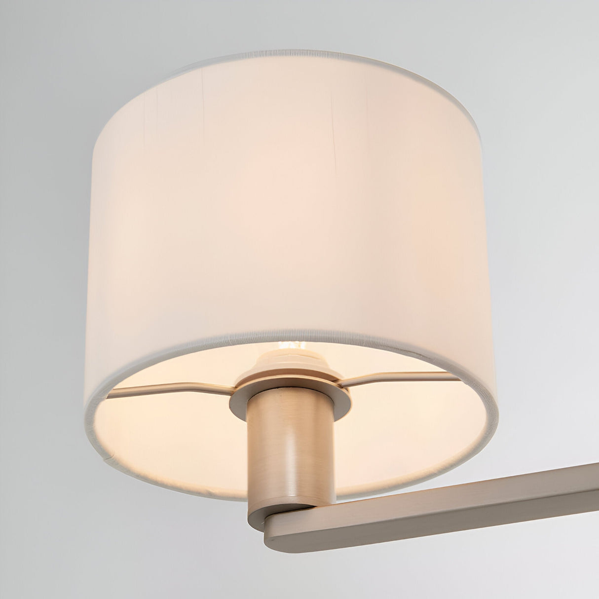 Bexley 3 Light Pendant in matt nickel, close-up of one white faux silk drum shade with lit bulb, arm and central fitting visible, plain light grey background.