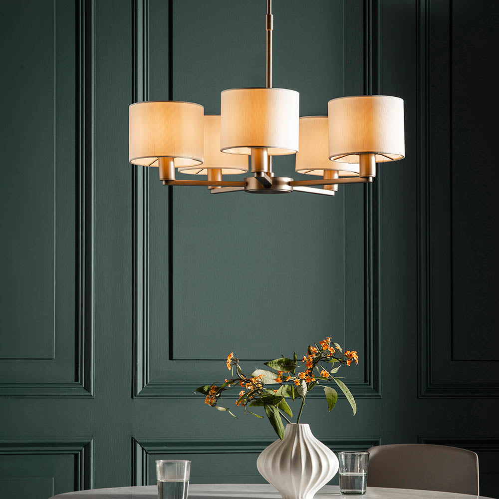 Bexley 5 Light Pendant in matt nickel with five curved arms and cream faux silk drum shades, hung above a dining table in a dark green panelled room.