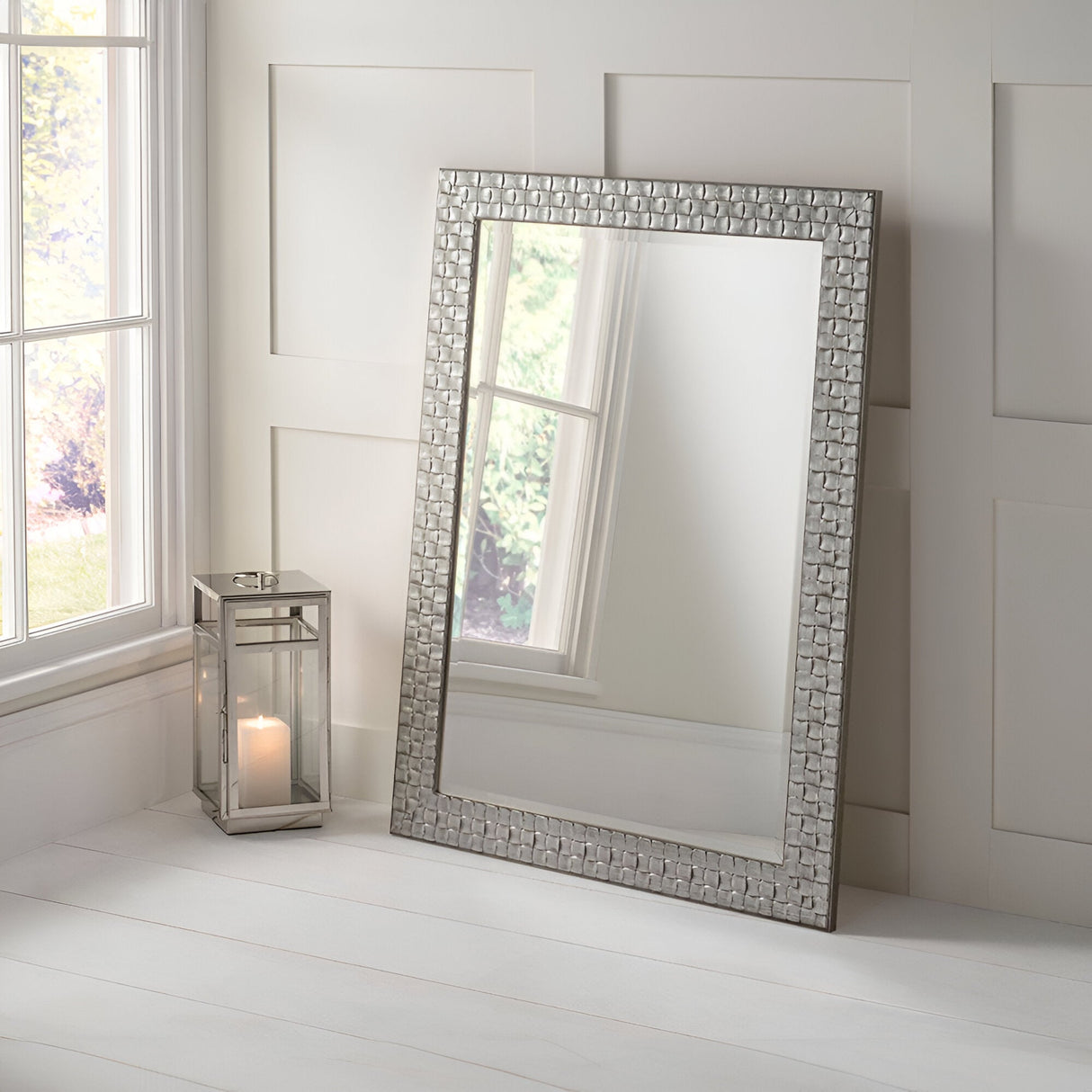 The Reflect Rectangle Wall Mirror - Silver, featuring an ornate frame, leans against a white paneled wall. Adjacent to it on the white wooden floor is a metallic lantern with a lit candle inside. Light from a window on the left highlights its modern design.