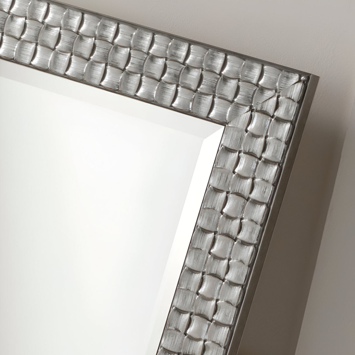 A close-up view of the Reflect Rectangle Wall Mirror - Silver showcases its textured, metallic frame with a basketweave pattern. The reflective silver frame elevates the modern design, adding an elegant touch as the mirror sits angled against a plain, light-colored wall.