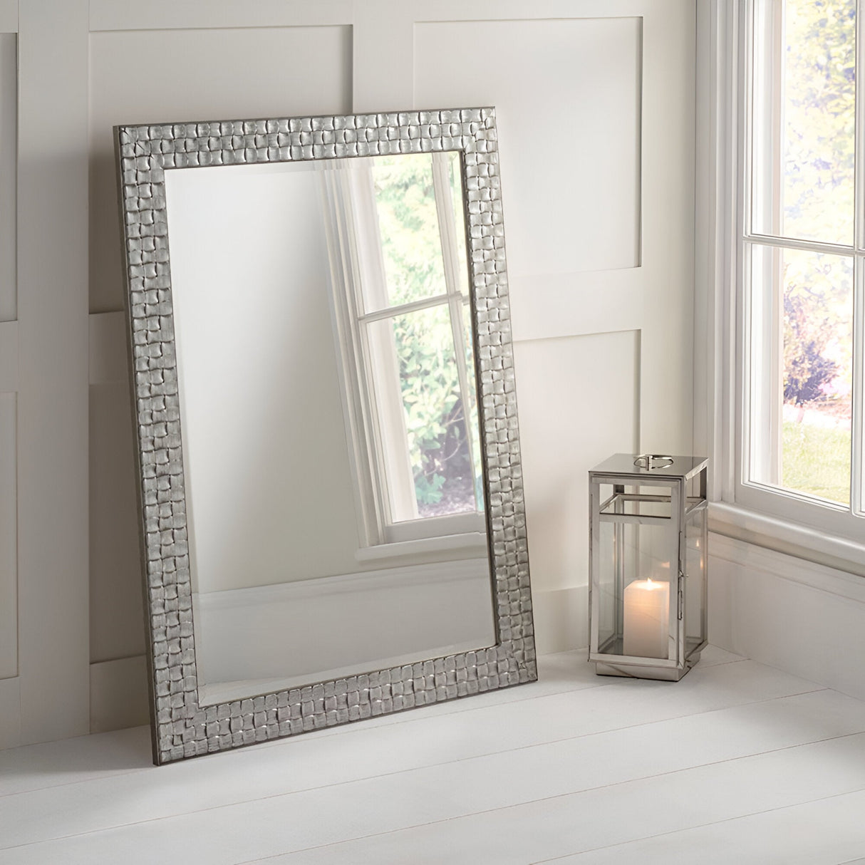 The Miraggio Leaner Mirror - White & Silver, with its ornate frame, leans against a light paneled wall beside a lit candle in a glass lantern. Positioned near a window with soft natural light streaming in, it creates an elegant decorative statement.