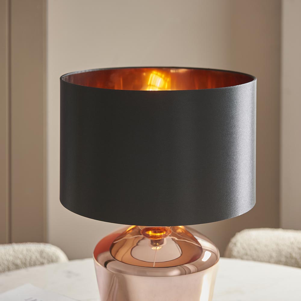 Endon Danefield table lamp with a polished copper glass base, black drum shade with copper-effect inner, and lit E27 bulb, on a marble table.