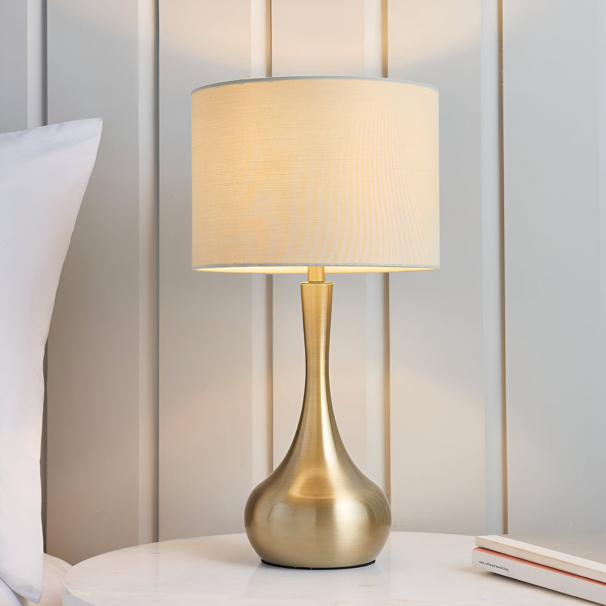 Endon Chartbury LED table lamp with a brass teardrop base and beige drum shade, lit on a bedside table beside a pillow and panelled wall.