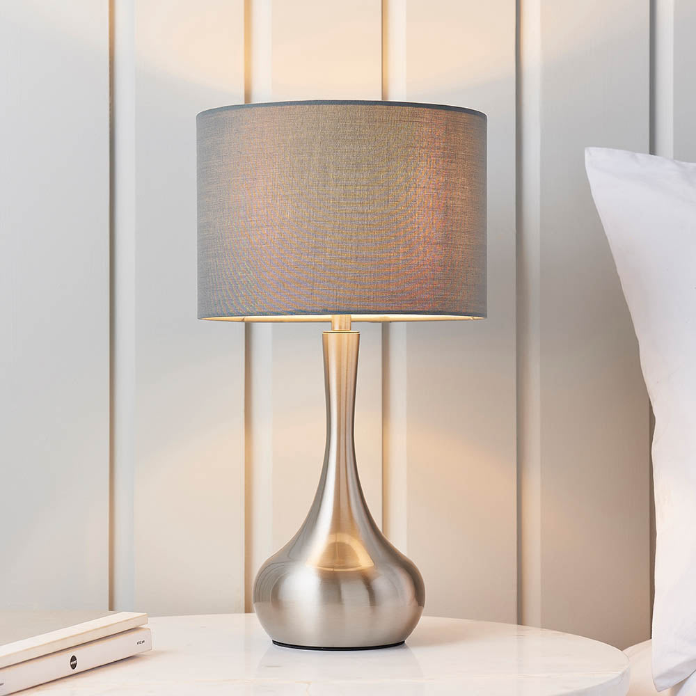 Endon Chartbury LED table lamp with satin nickel curved base and grey fabric drum shade, lit on a marble bedside table beside a pillow.
