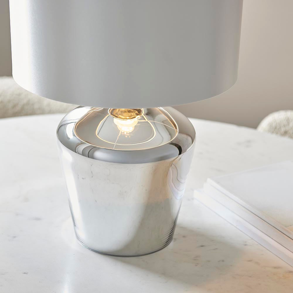 Danefield Table Lamp in chrome, close-up of the rounded glass base with lit E27 bulb beneath a cream fabric shade, set on a marble table.