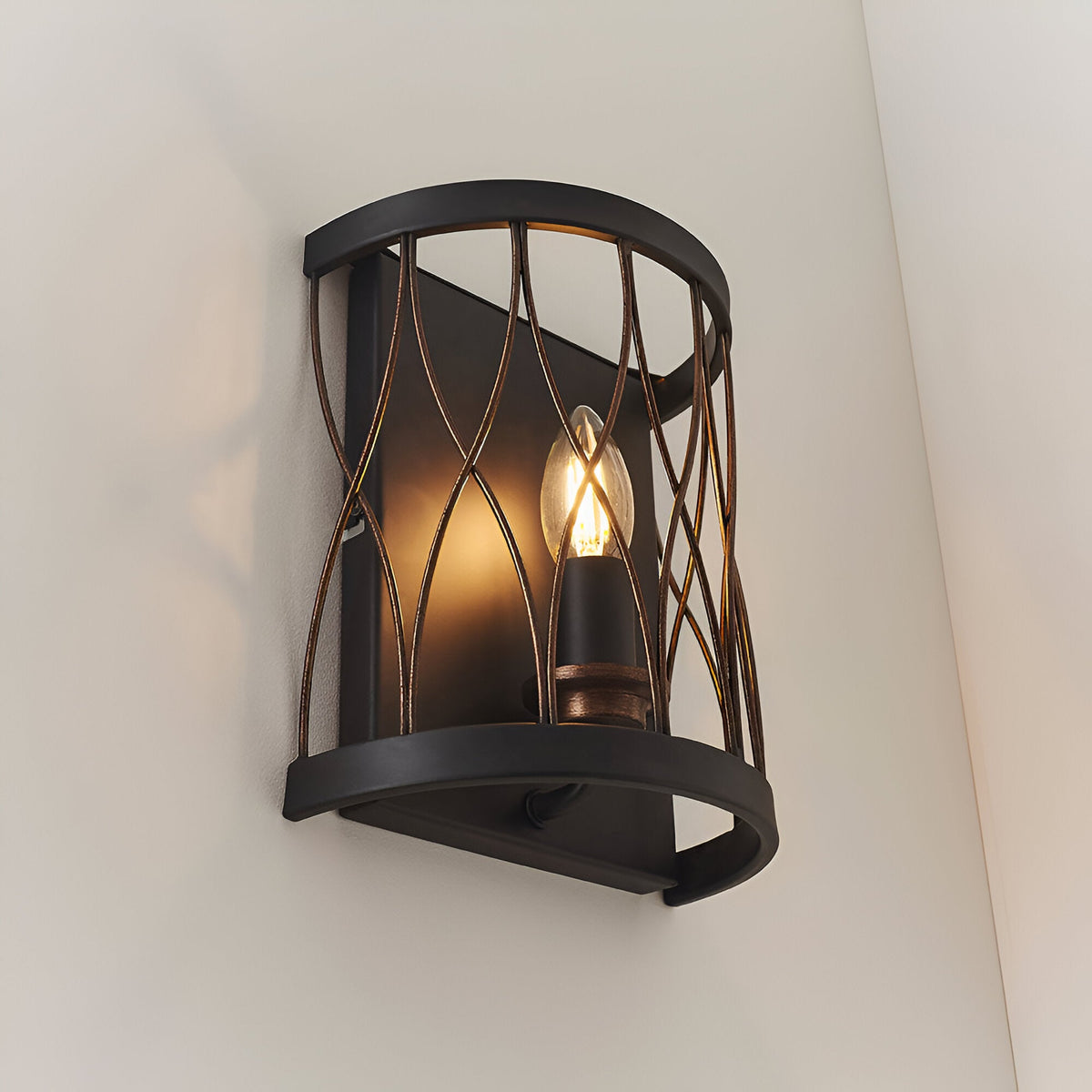 Endon Hulton wall light with a curved steel cage in matt black and rustic bronze, illuminated E14 candle bulb, mounted on a pale corner wall.