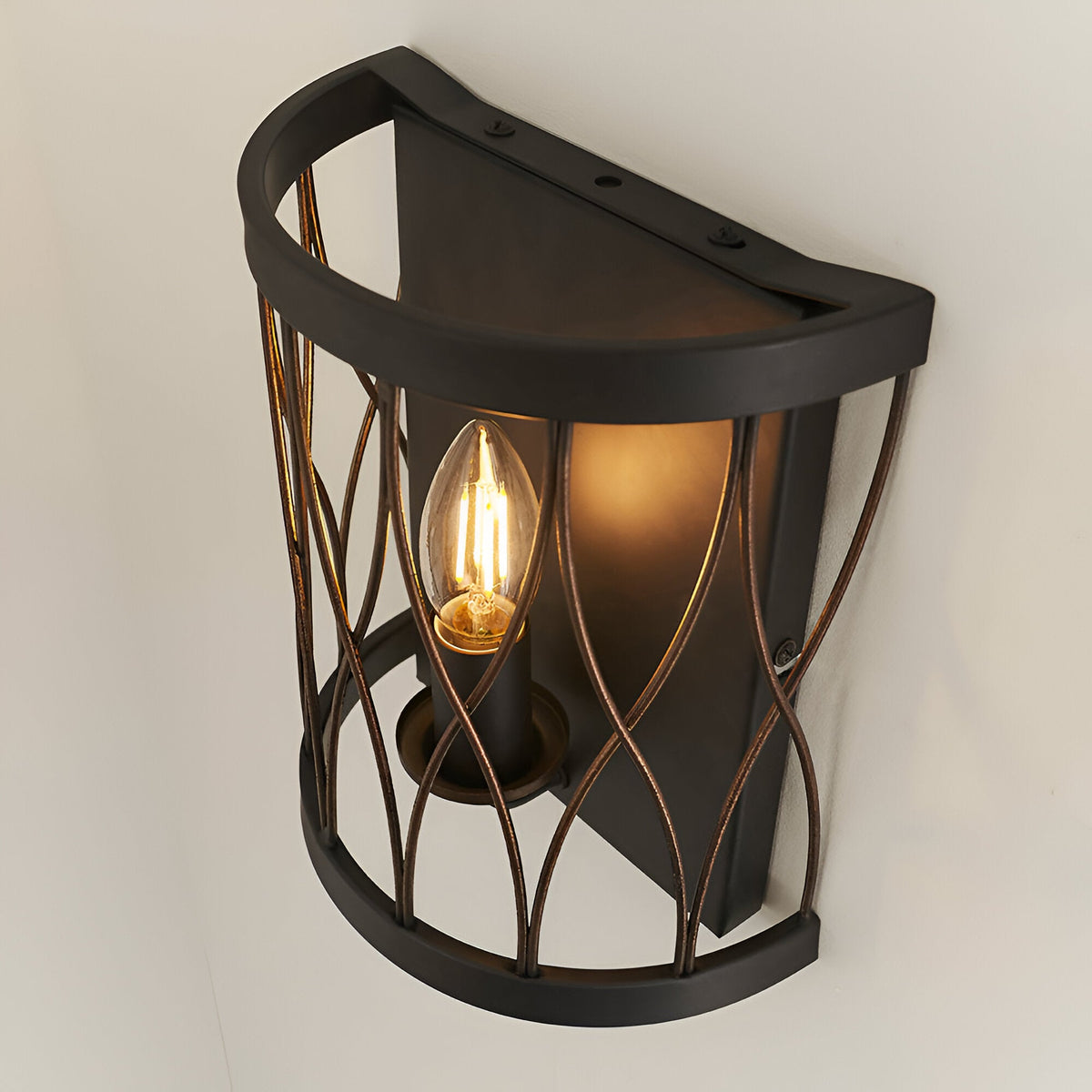 Endon Hulton wall light with a curved steel cage in matt black and rustic bronze, single lit E14 candle bulb, mounted on a white wall.