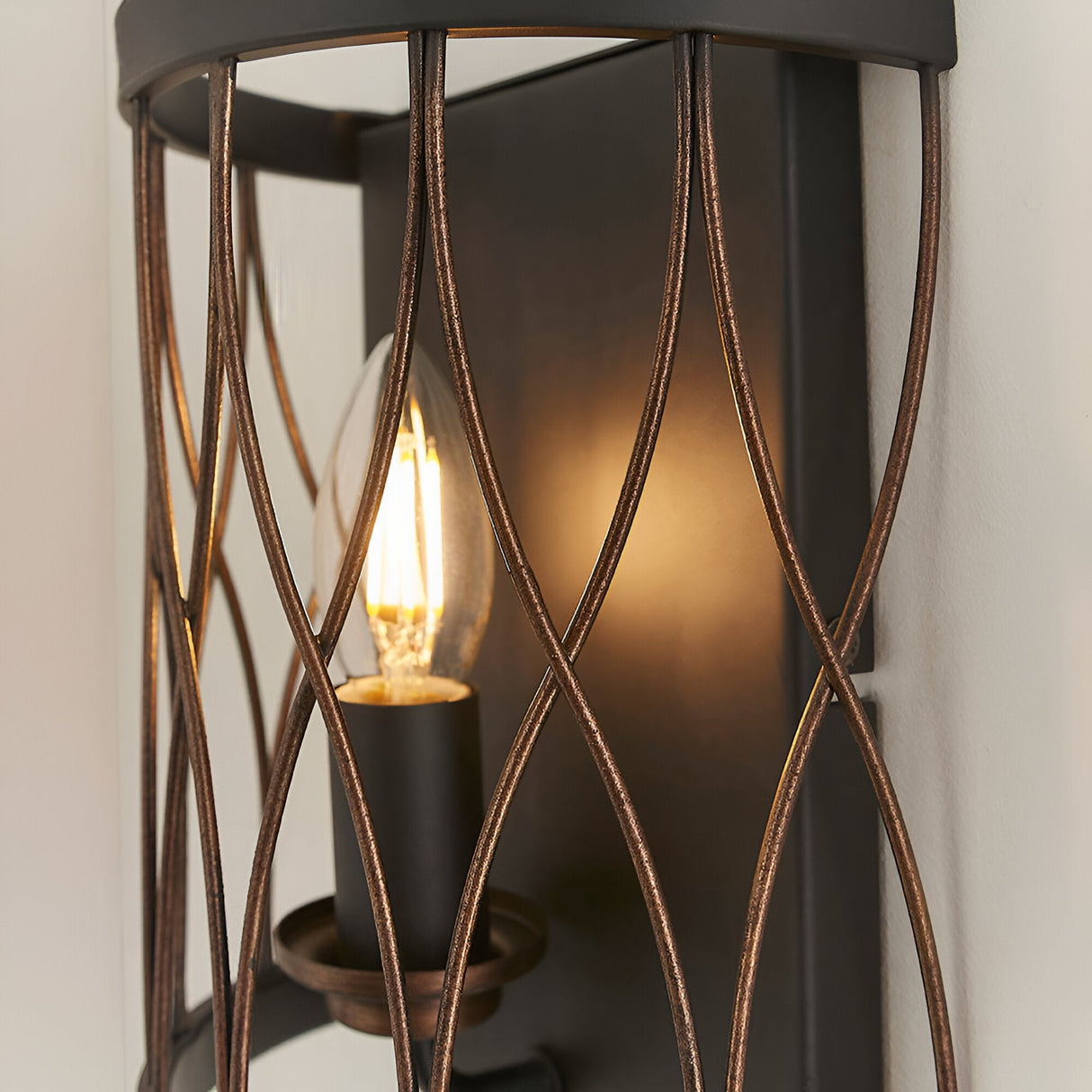 Close-up of the Endon Hulton wall light showing its crossed bronze wirework cage, matt black backplate and lit E14 candle bulb.