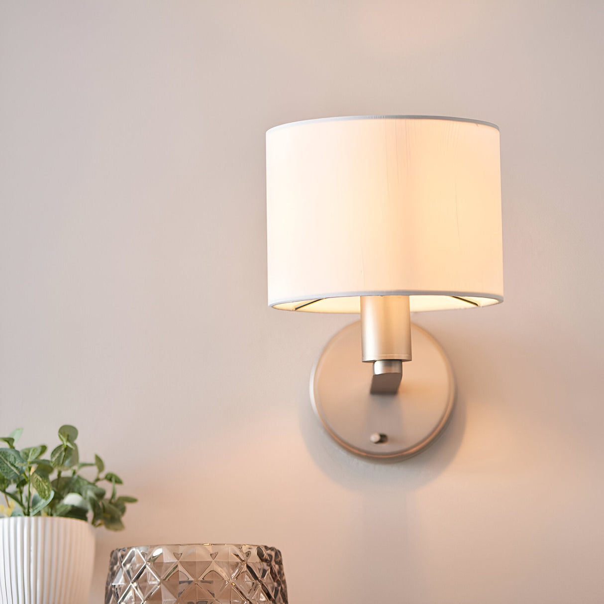 Bexley Wall Light in nickel with a white faux silk drum shade, shown illuminated on a light wall above a plant and glass vase.