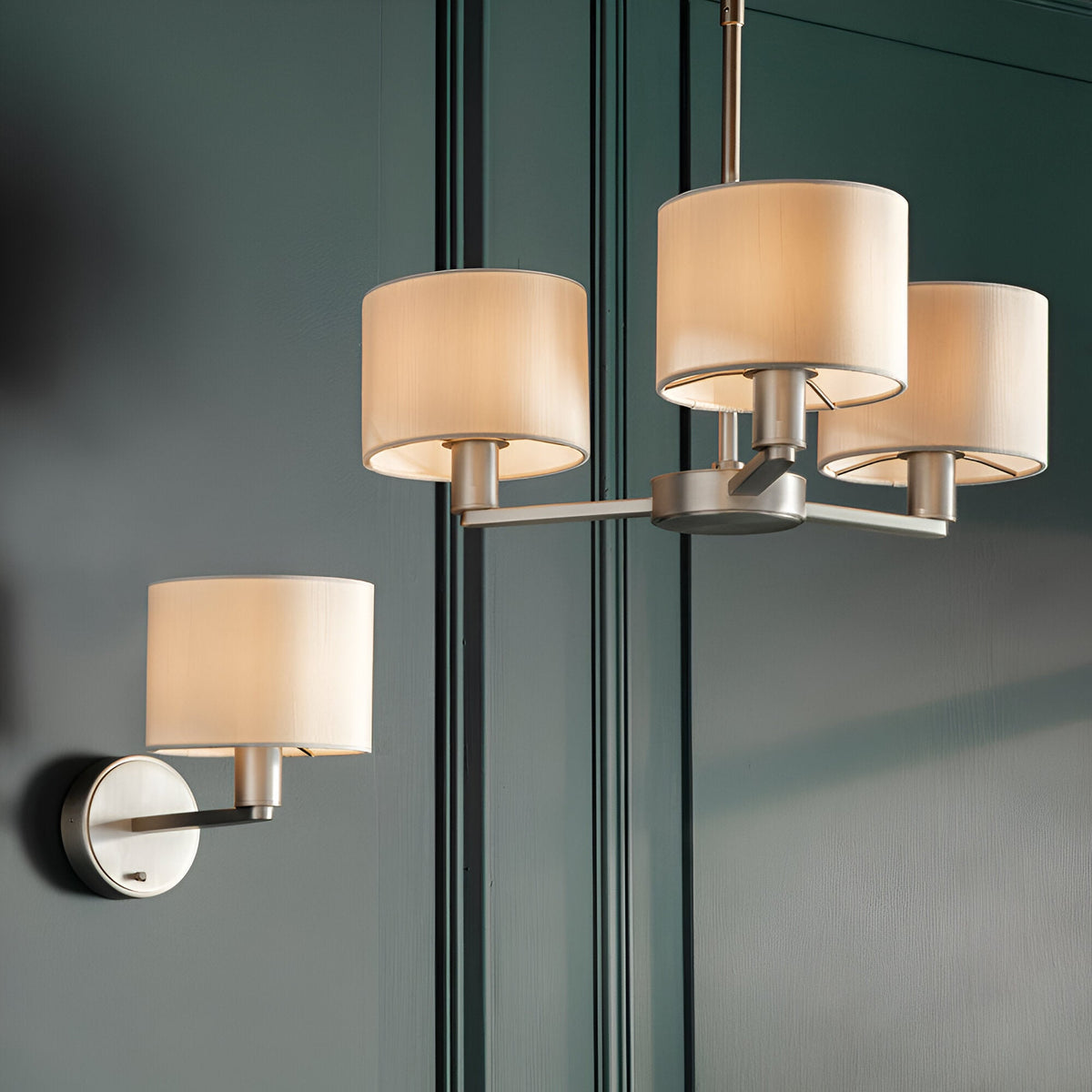Bexley Wall Light in nickel with white drum shade, mounted on a dark green panelled wall alongside a matching chandelier.