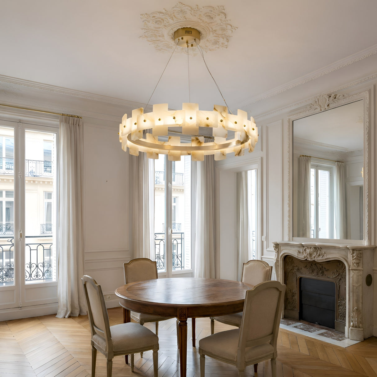A round wooden dining table with four beige chairs sits in an elegant room with large windows, a decorative fireplace, a large mirror, and the Ravion LED Small Panelled Alabaster Chandelier - Soft White & Brass hanging from the ceiling.