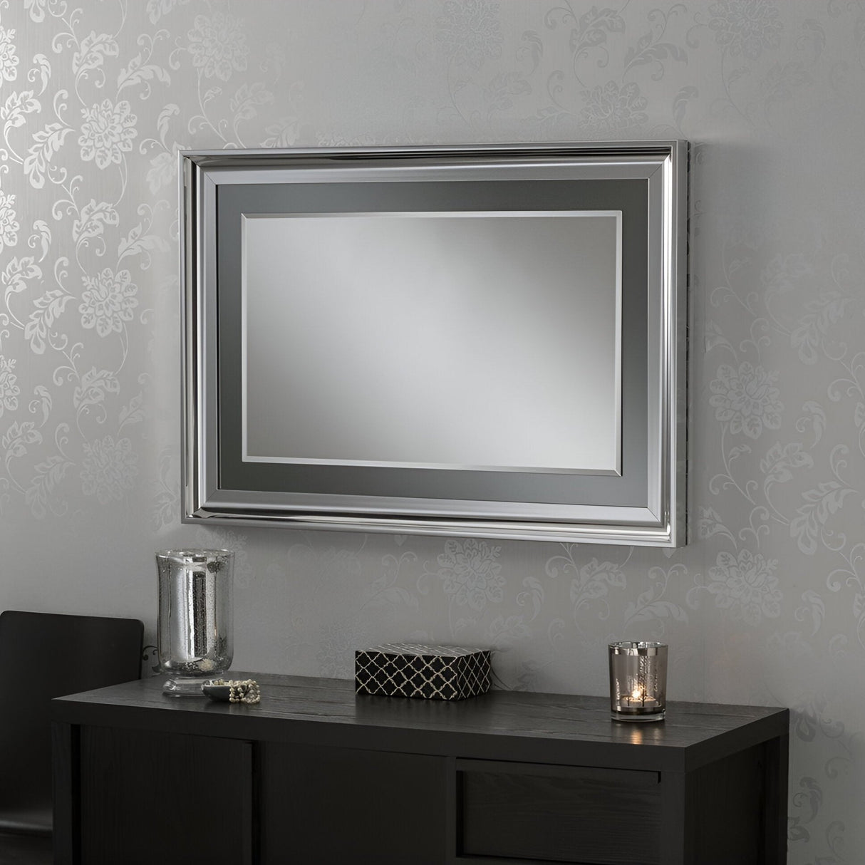 A Wave Rectangle Wall Mirror with a silver lacquered finish elegantly frames the view of floral patterned wallpaper. Below it, a dark wooden console table is adorned with tasteful decorative items like a candle and vase, creating an elegant ambiance.