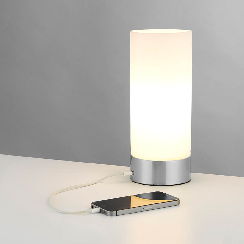 Endon Bracewell nickel table lamp with brushed metal base, matt opal glass cylindrical shade and USB port, shown charging a smartphone on a light surface.