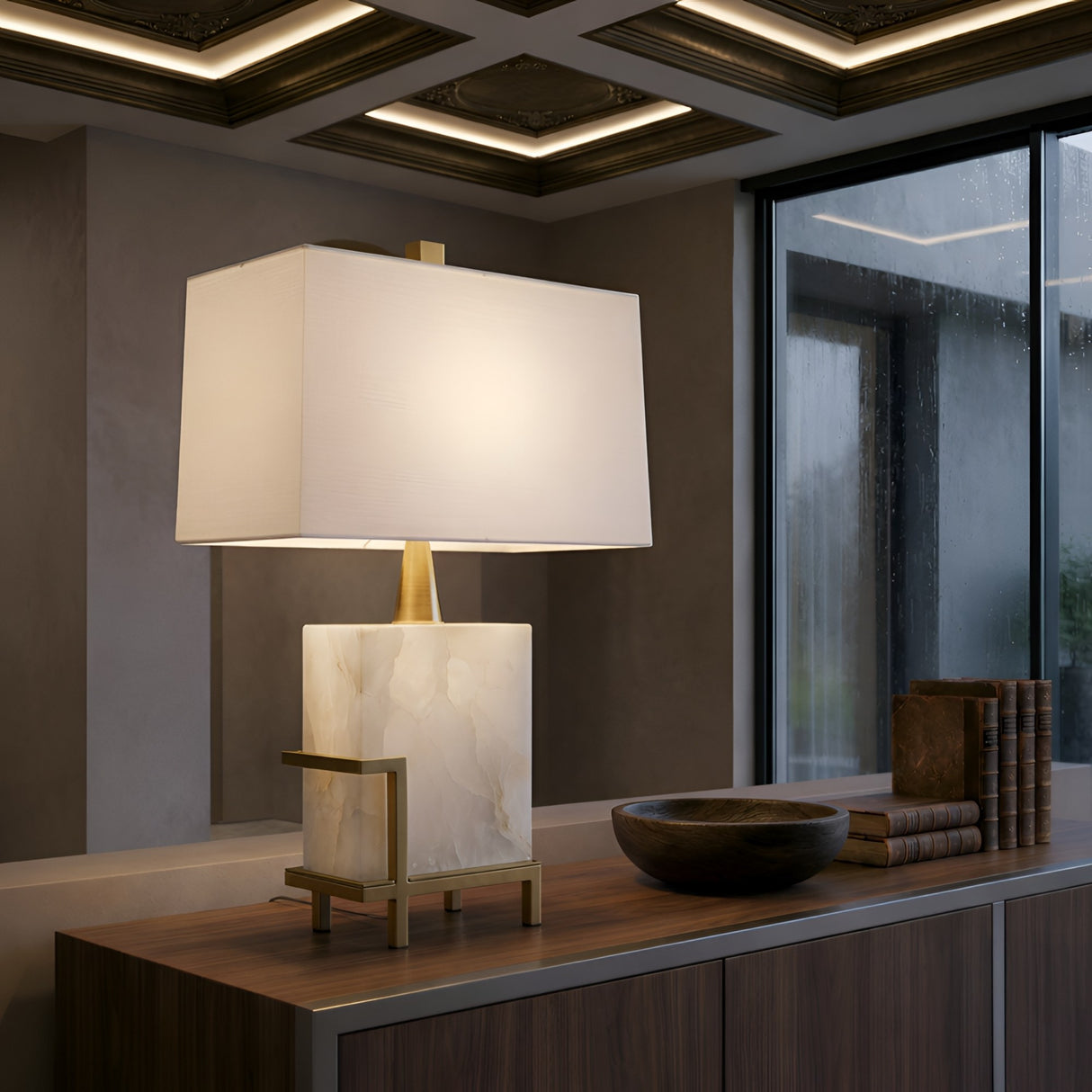 The Lumenis 1 Light Alabaster Table Lamp – Brass & Soft White sits on a wooden sideboard beside a stack of books and a wooden bowl, near large windows overlooking rain.