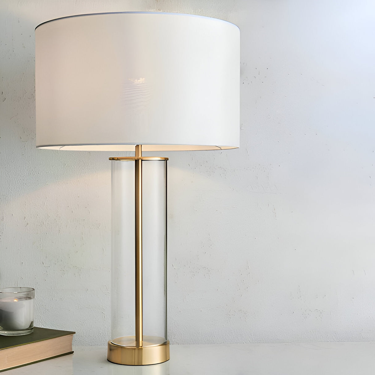 Endon Caldley table lamp with clear cylindrical glass base, brass fittings, and vintage white drum shade, styled on a side table beside a candle.