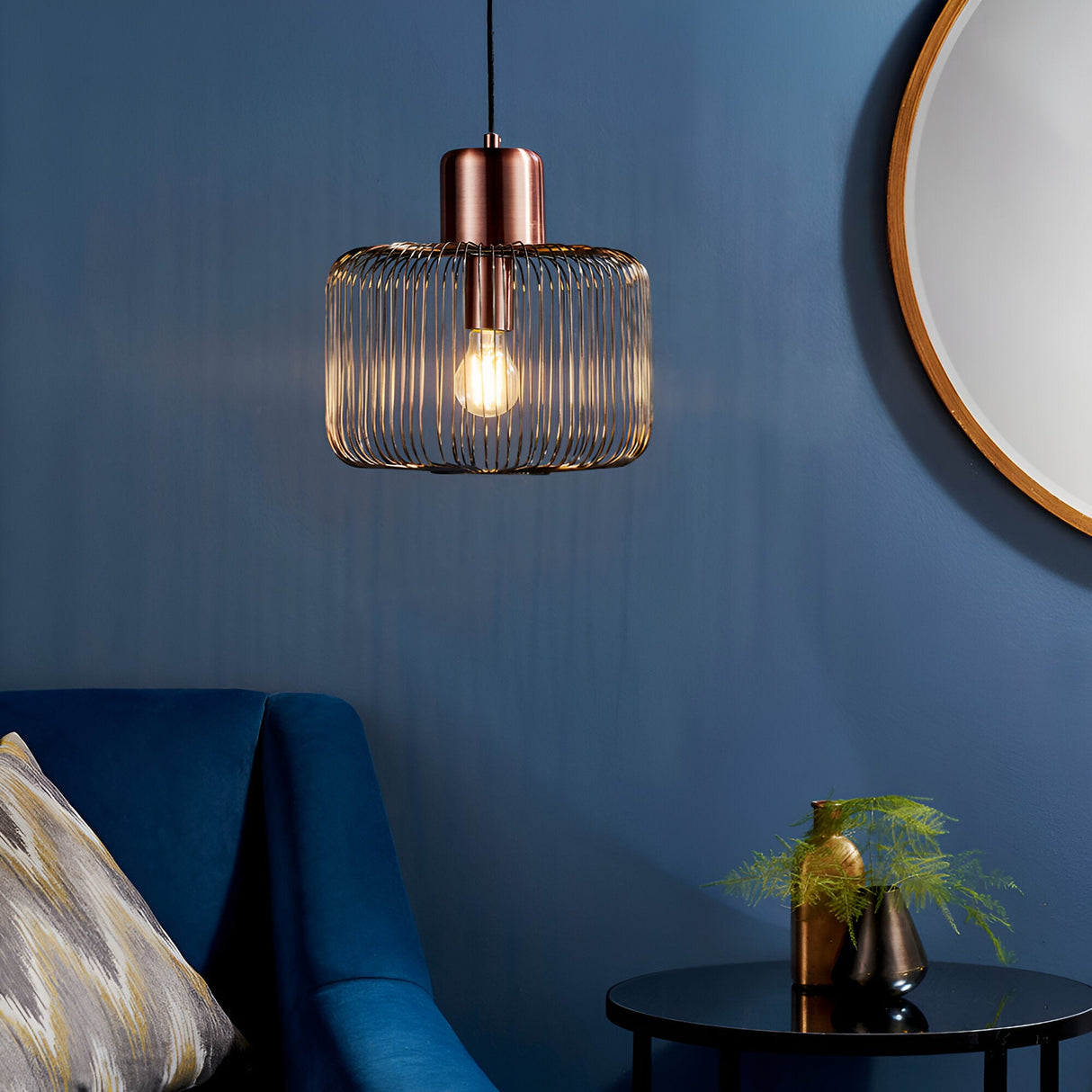 Castdale Cage Pendant in antique copper with open wire cage shade and exposed bulb, hung above a blue sofa near a round mirror.