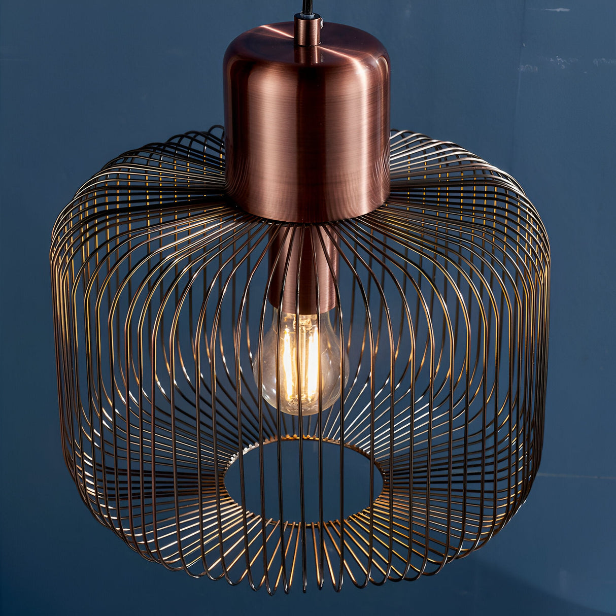 Castdale Cage Pendant in antique copper finish, open wire cage shade with visible bulb, shown against a dark blue background.