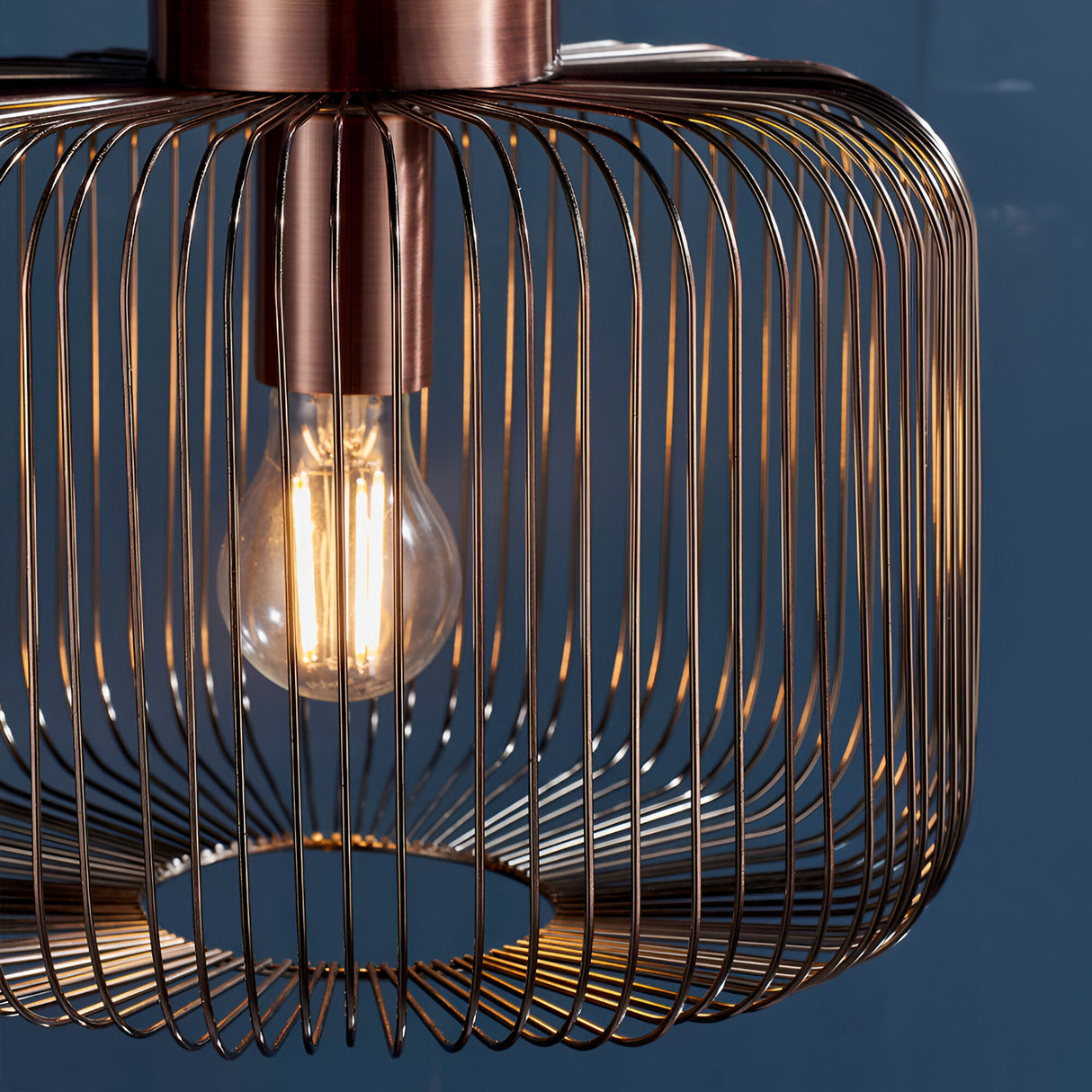 Castdale Cage Pendant in antique copper finish, close-up of open wire cage shade with visible E27 bulb, set against a dark blue background.