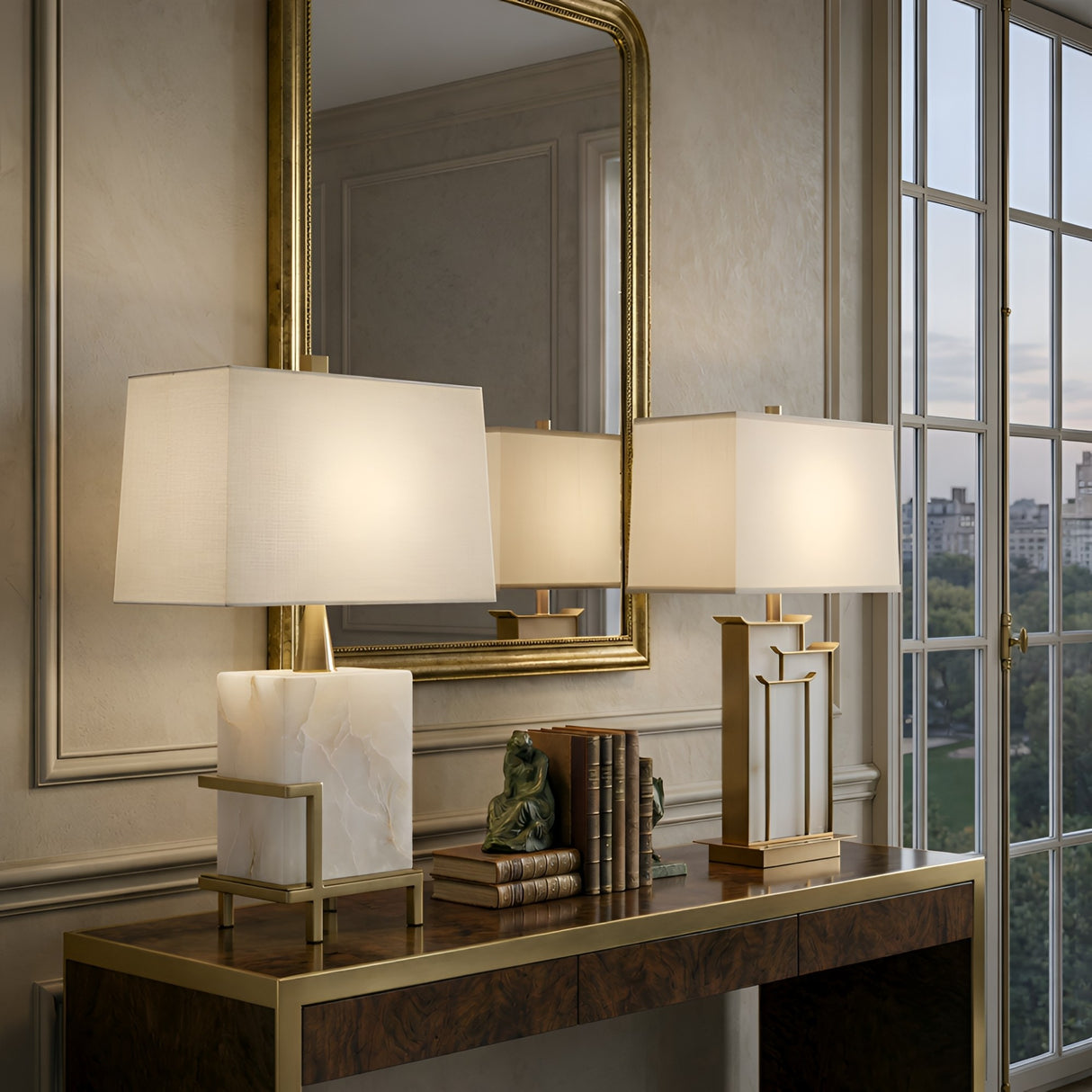 A brass console table styled with two Veliora 1 Light Framed Alabaster Table Lamps in Brass & Soft White, books, and a small sculpture sits before a large mirror and tall window with city views, creating an elegant, sophisticated setting.