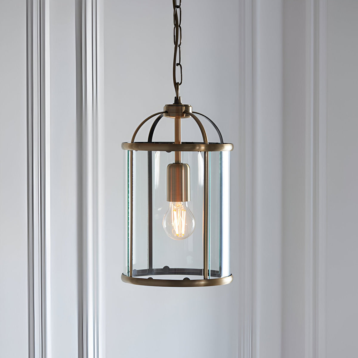 Caldbury Pendant Light in brass, cylindrical lantern frame with clear glass panels and exposed E27 bulb, hung from a chain against a plain white wall.