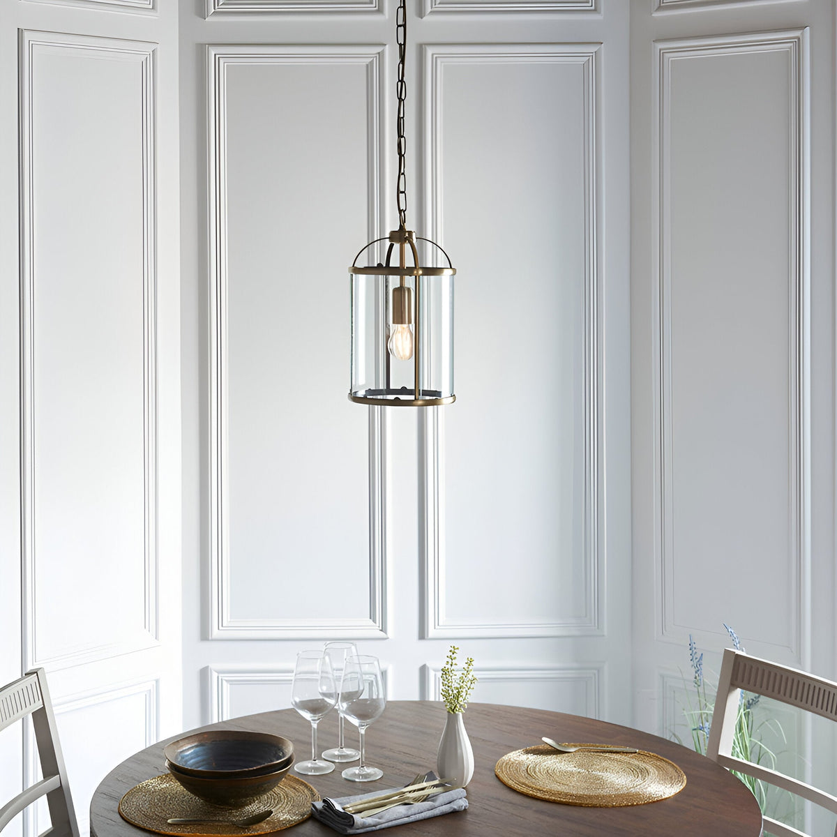 Caldbury Pendant Light in brass, single E27 lantern-style fixture with clear glass panels, hung above a dining table in a panelled white room.