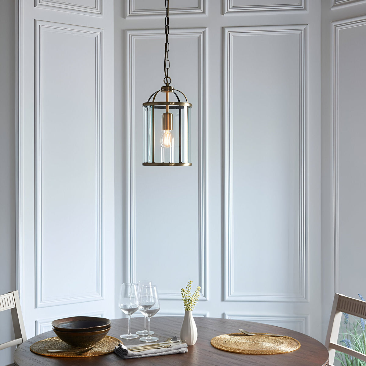 Caldbury Pendant Light in brass, a single lantern-style fixture with clear glass panels and exposed bulb, hung from a chain above a dining table.