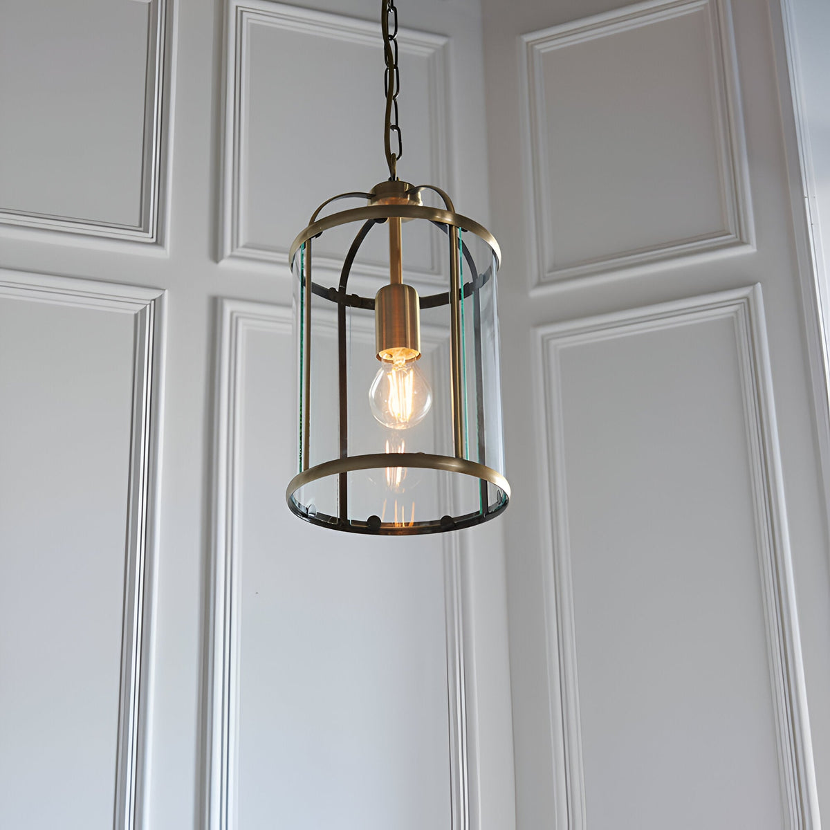 Caldbury Pendant Light in brass, a cylindrical lantern-style fixture with clear glass panels and a visible filament bulb, hung from a chain.