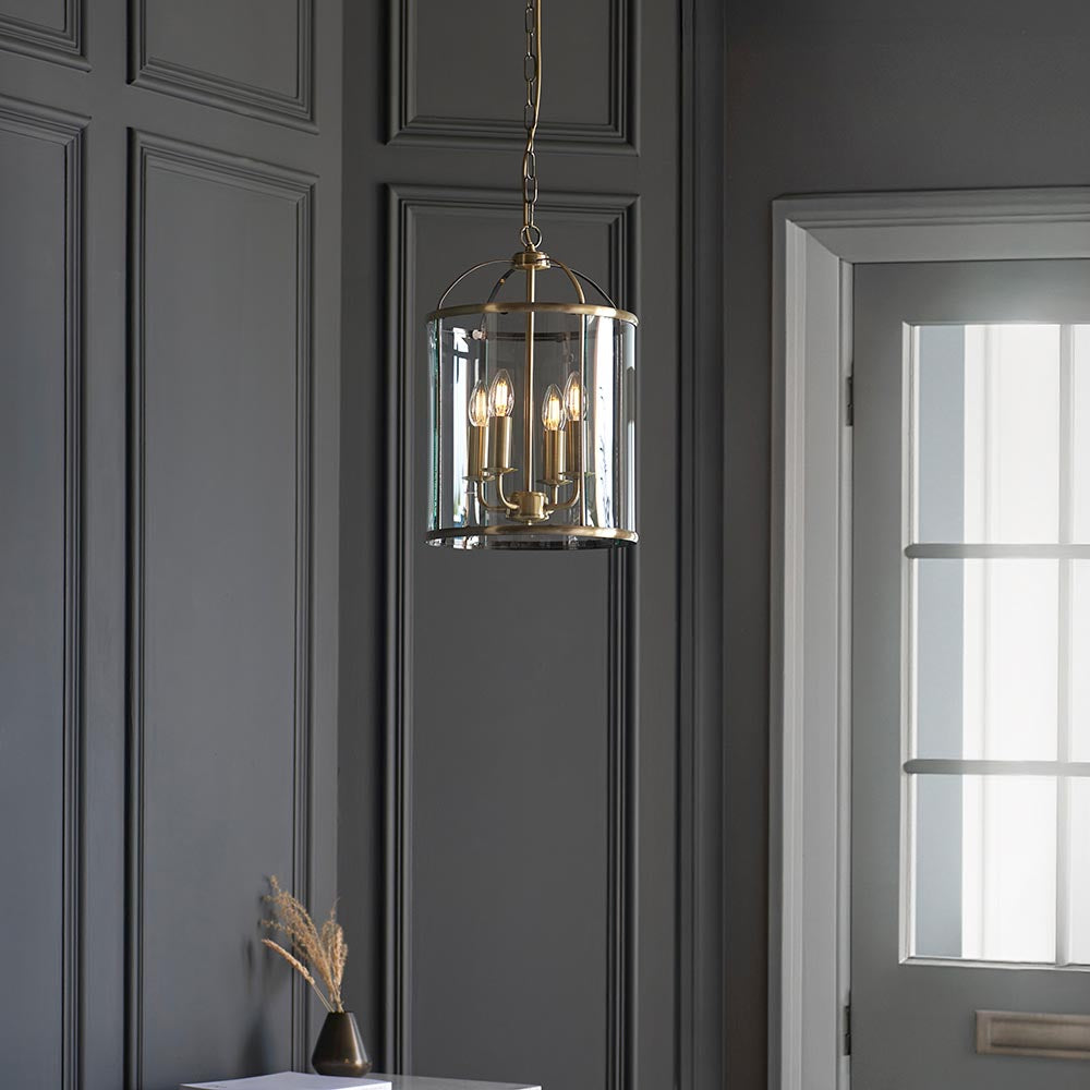 Caldbury 4 Light Pendant in brass with clear glass panels and candle-style bulbs, hanging on a chain against dark panelled walls.