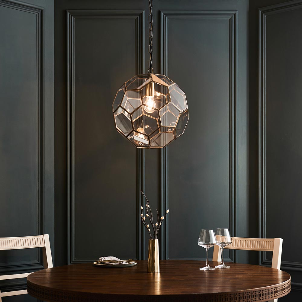 Endon Carldon Hexagonal Pendant in antique brass with clear glass panels, suspended by chain above a round wooden dining table against a dark panelled wall.