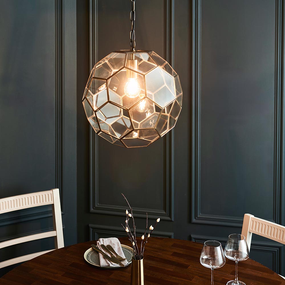 Endon Carldon hexagonal glass pendant in antique brass, illuminated over a wooden dining table against a dark panelled wall.