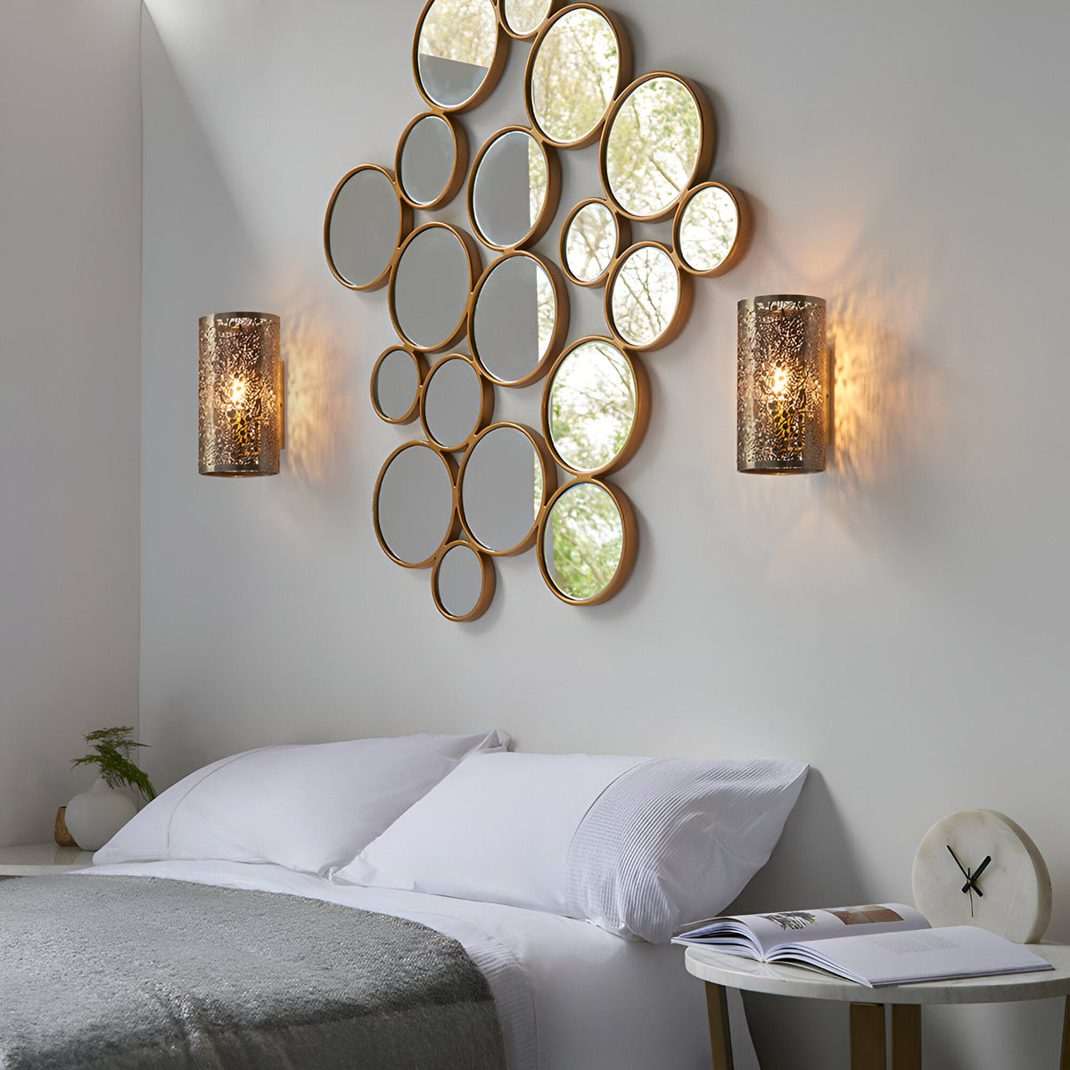 Pair of Endon Cliffmere antique brass wall lights flanking a circular mirror collage above a bed, casting patterned light across the wall.