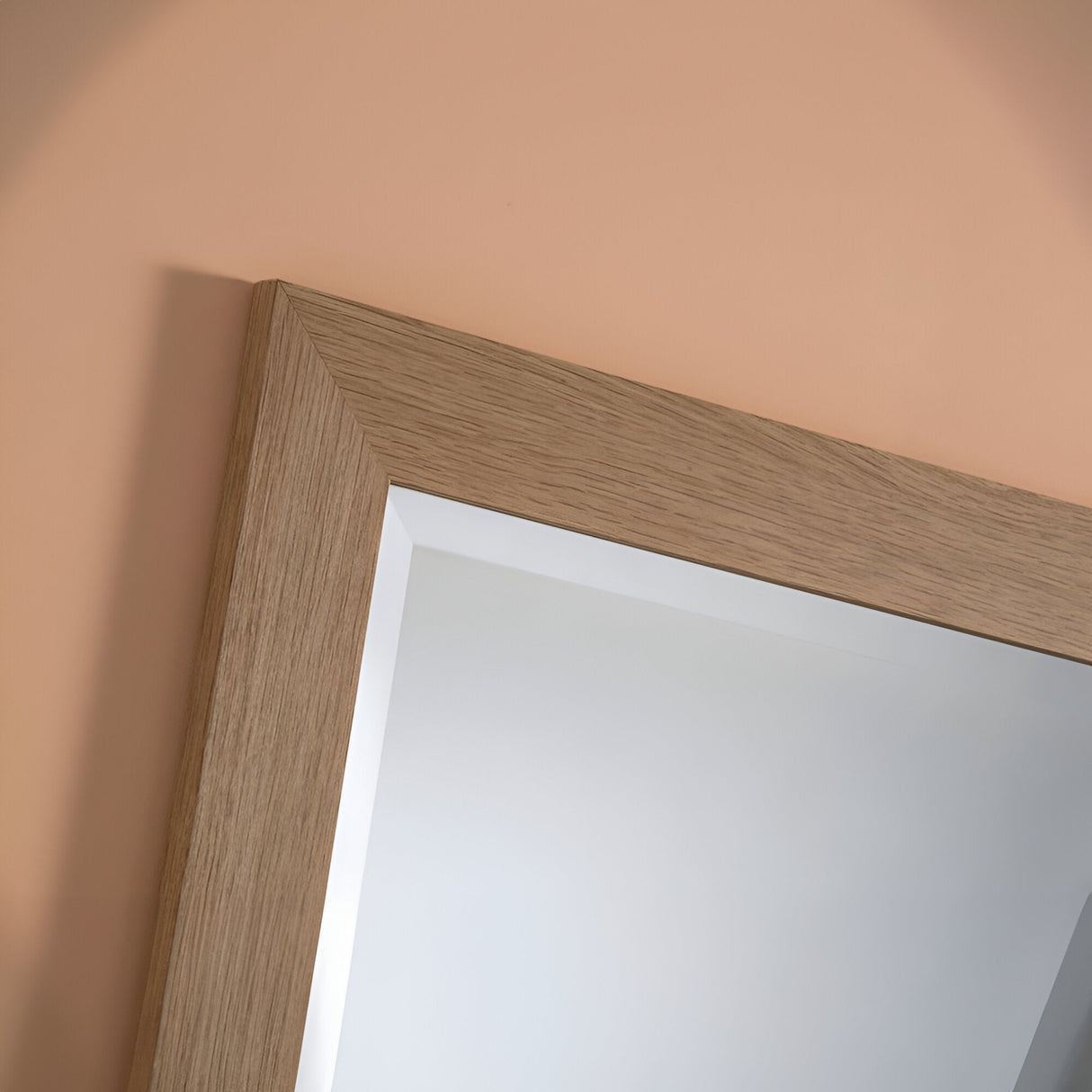 The Vestige Rectangle Wall Mirror - Oak is showcased up-close, hanging on a beige wall. Its oak frame highlights the natural wood grain texture, adding rustic charm that contrasts beautifully with the smooth mirror surface. The reflection in the mirror reveals a pristine white ceiling.