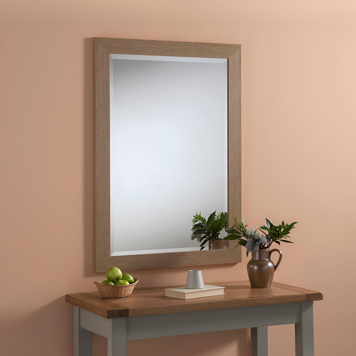 A simple wooden table exudes rustic charm with a decorative vase of greenery and flowers on the right, and a green bowl filled with apples on the left. Above it, the beautifully crafted Vestige Rectangle Wall Mirror - Oak elegantly hangs against the peach-colored wall.